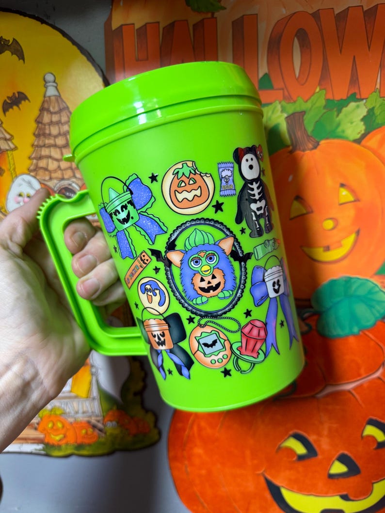 Puede incluir: Una taza de viaje de pl&aacute;stico de color verde lima brillante con asa y tapa, que presenta un estampado colorido alrededor con varios iconos tem&aacute;ticos de Halloween, incluyendo un personaje Furby central, calabazas y lazos.