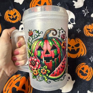 May include: A clear, glittery plastic mug with a handle featuring a colorful graphic of a jack-o'-lantern carved from a watermelon, surrounded by vibrant flowers and a slice of watermelon. The mug has a screw-top lid and a textured, translucent finish.