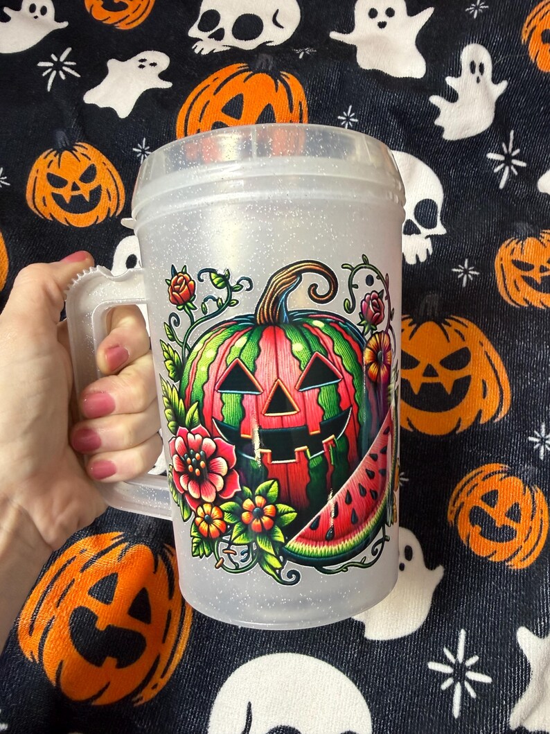 May include: A clear, glittery plastic mug with a handle featuring a colorful graphic of a jack-o'-lantern carved from a watermelon, surrounded by vibrant flowers and a slice of watermelon. The mug has a screw-top lid and a textured, translucent finish.