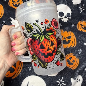 May include: A clear, glittery plastic mug with a handle, featuring a graphic illustration of a red strawberry designed with a jack-o'-lantern face, surrounded by green leaves, small red berries, and red flowers on the front surface.