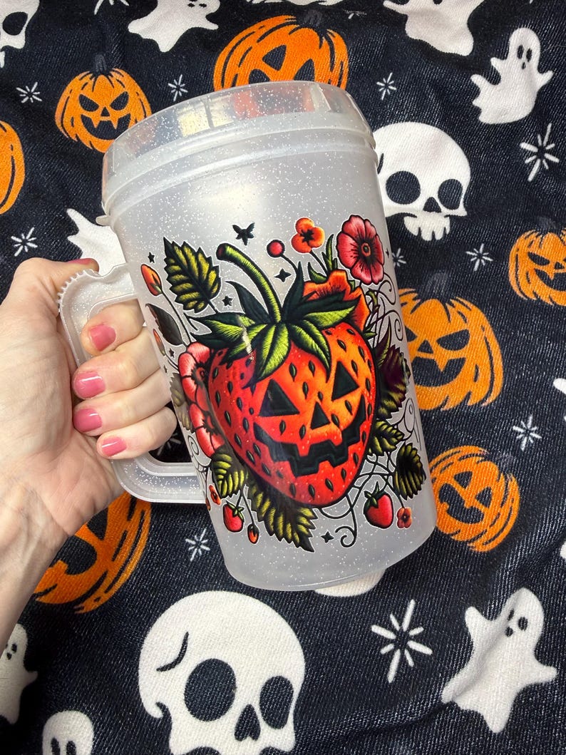 May include: A clear, glittery plastic mug with a handle, featuring a graphic illustration of a red strawberry designed with a jack-o'-lantern face, surrounded by green leaves, small red berries, and red flowers on the front surface.