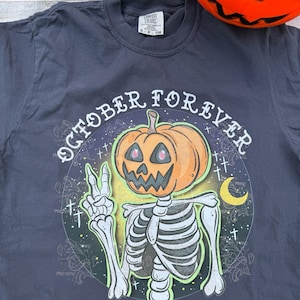 Peut inclure: T-shirt gris foncé avec un motif sur le thème d'Halloween. Le motif représente un squelette avec une tête de citrouille, faisant le signe de la paix, et les mots "OCTOBER FOREVER" dans un cercle.