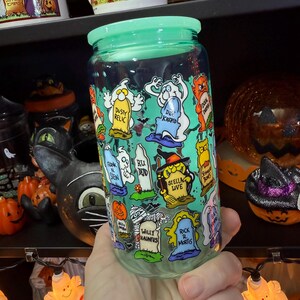 Peut inclure: Un bocal en verre transparent avec un couvercle vert clair, d&eacute;cor&eacute; de pierres tombales et de fant&ocirc;mes de dessins anim&eacute;s. Le bocal comporte des phrases comme "Dusty Relic", "Dee Krepid" et "Willy Hauntus". Le bocal est tenu dans une main, avec des d&eacute;corations d'Halloween en arri&egrave;re-plan.