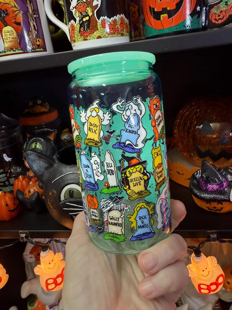 Peut inclure: Un bocal en verre transparent avec un couvercle vert clair, d&eacute;cor&eacute; de pierres tombales et de fant&ocirc;mes de dessins anim&eacute;s. Le bocal comporte des phrases comme "Dusty Relic", "Dee Krepid" et "Willy Hauntus". Le bocal est tenu dans une main, avec des d&eacute;corations d'Halloween en arri&egrave;re-plan.