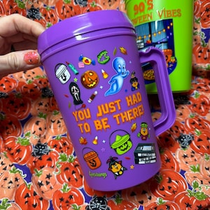 Op de afbeelding: Een paarse plastic beker met handvat voorzien van een Casper het vriendelijke spookje-afbeelding, diverse Halloween-iconen en de tekst "YOU JUST HAD TO BE THERE!" in het oranje. Een groene beker is gedeeltelijk zichtbaar op de achtergrond.
