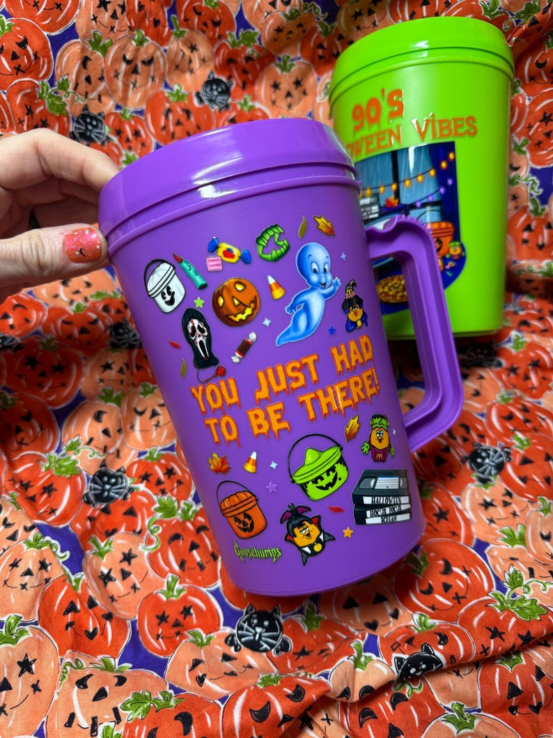 Op de afbeelding: Een paarse plastic beker met handvat voorzien van een Casper het vriendelijke spookje-afbeelding, diverse Halloween-iconen en de tekst "YOU JUST HAD TO BE THERE!" in het oranje. Een groene beker is gedeeltelijk zichtbaar op de achtergrond.