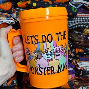 May include: A bright orange plastic mug with a handle, featuring a colorful graphic of cartoon monsters and the black text "LETS DO THE MONSTER MASH" printed on the front. The mug has a matching orange lid and a smooth, cylindrical body design.
