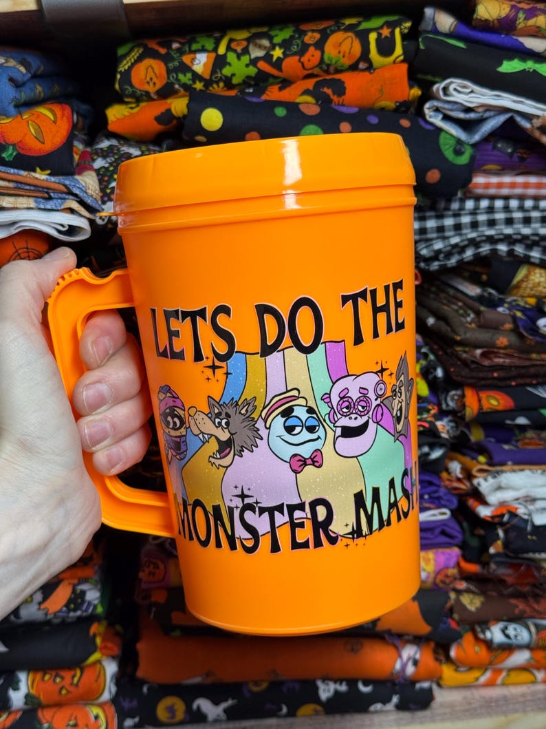 May include: A bright orange plastic mug with a handle, featuring a colorful graphic of cartoon monsters and the black text "LETS DO THE MONSTER MASH" printed on the front. The mug has a matching orange lid and a smooth, cylindrical body design.