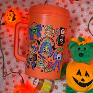 Puede incluir: Una taza de viaje de pl&aacute;stico naranja con asa, decorada con varias pegatinas coloridas con tem&aacute;tica de Halloween que incluyen un Furby central, calabazas, lazos, un oso esqueleto y un chupete, sobre un fondo de luces de calabaza naranjas.