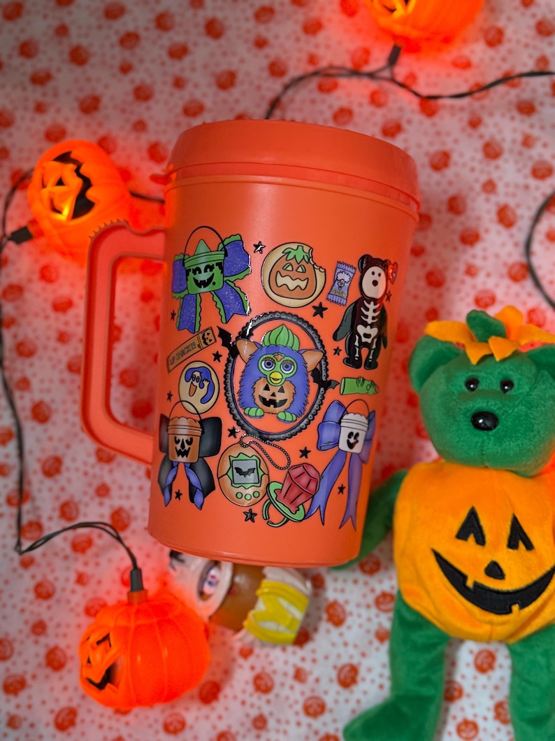 Puede incluir: Una taza de viaje de pl&aacute;stico naranja con asa, decorada con varias pegatinas coloridas con tem&aacute;tica de Halloween que incluyen un Furby central, calabazas, lazos, un oso esqueleto y un chupete, sobre un fondo de luces de calabaza naranjas.