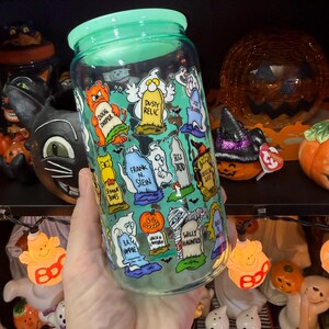 Peut inclure: Un bocal en verre transparent avec un couvercle vert, d&eacute;cor&eacute; de personnages de dessins anim&eacute;s et de pierres tombales sur le th&egrave;me d'Halloween. Le bocal pr&eacute;sente des images de fant&ocirc;mes, de citrouilles et d'autres &eacute;l&eacute;ments effrayants. Le texte sur le bocal comprend des phrases comme "Doug Deeper" et "Willy Hauntus".