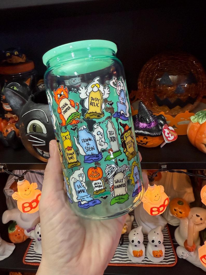 Peut inclure: Un bocal en verre transparent avec un couvercle vert, d&eacute;cor&eacute; de personnages de dessins anim&eacute;s et de pierres tombales sur le th&egrave;me d'Halloween. Le bocal pr&eacute;sente des images de fant&ocirc;mes, de citrouilles et d'autres &eacute;l&eacute;ments effrayants. Le texte sur le bocal comprend des phrases comme "Doug Deeper" et "Willy Hauntus".