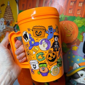以下が含まれることがあります： オレンジ色のプラスチック製ハロウィーンマグカップ。ハンドルと蓋が付いています。マグカップは、カボチャ、幽霊、スケルトンなど、さまざまなハロウィーンをテーマにした漫画のキャラクターとシンボルで飾られています。マグカップは人の手で持たれています。