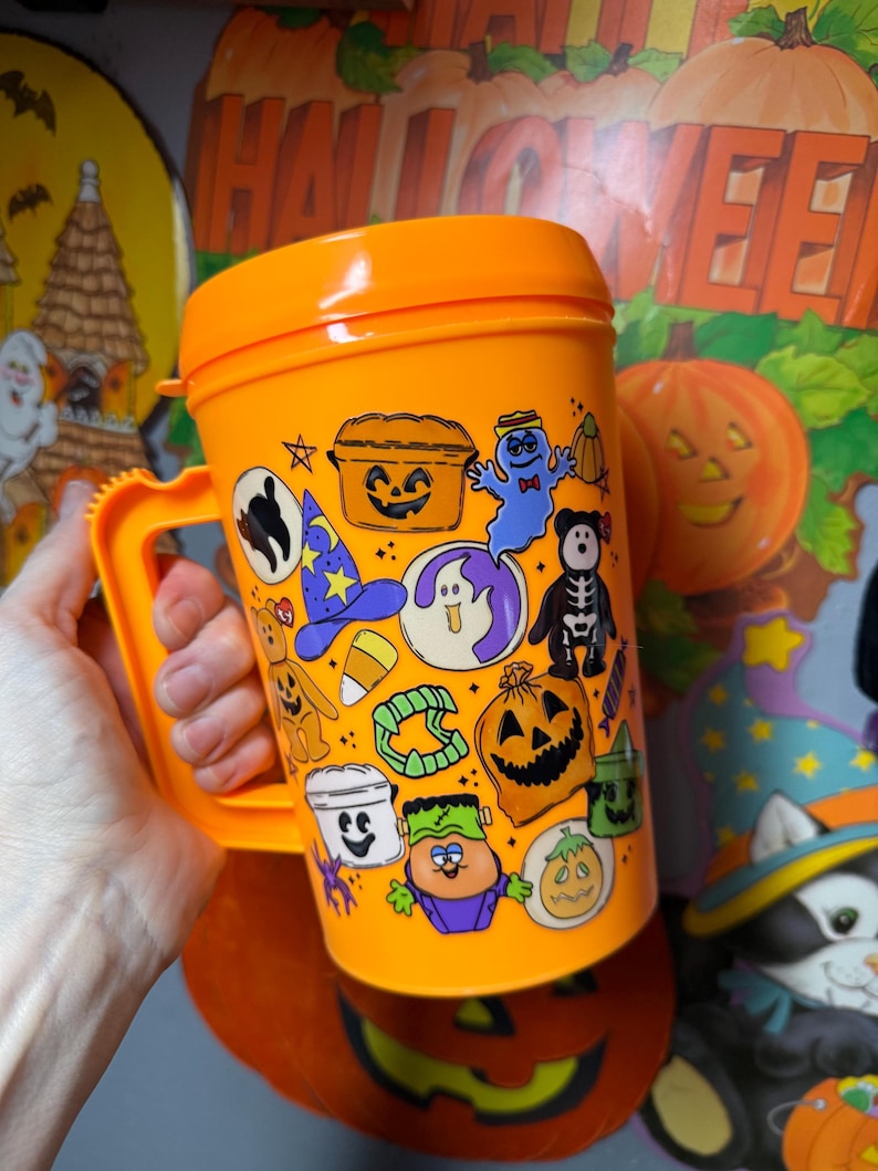 以下が含まれることがあります： オレンジ色のプラスチック製ハロウィーンマグカップ。ハンドルと蓋が付いています。マグカップは、カボチャ、幽霊、スケルトンなど、さまざまなハロウィーンをテーマにした漫画のキャラクターとシンボルで飾られています。マグカップは人の手で持たれています。