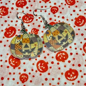 May include: A pair of dangle earrings featuring flat, pumpkin-shaped charms with a patchwork design. The pumpkins display various patterns including spiderwebs, stars, and dots in shades of orange, yellow, and purple, with silver-tone hooks.