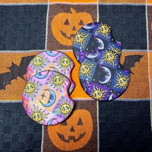 Peut inclure: Deux sous-verres circulaires pour porte-gobelet de voiture avec des motifs célestes. L'un présente un damier rose et orange avec des soleils jaunes et des croissants de lune. L'autre arbore un fond galaxie violet foncé et noir avec des soleils.