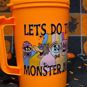 May include: An orange plastic mug with a lid and handle featuring a graphic of four cartoon monsters and the text LETS DO THE MONSTER MASH in black lettering. The design includes a mummy, a werewolf, a ghost, and a pink creature.
