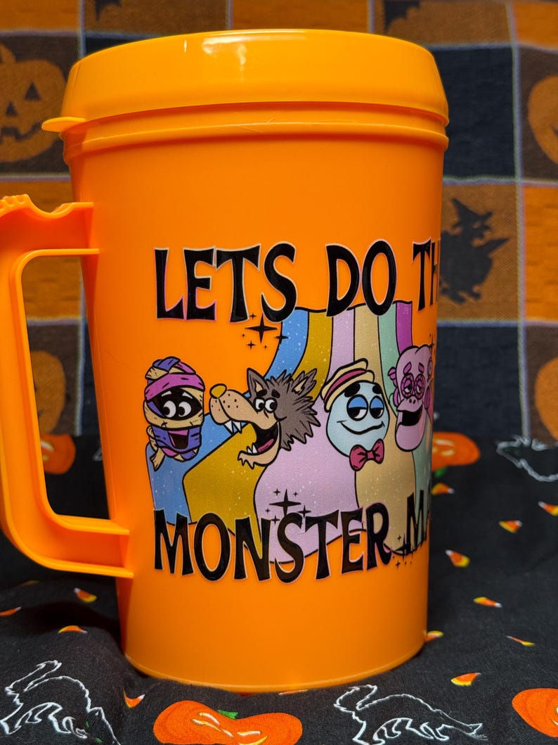 May include: An orange plastic mug with a lid and handle featuring a graphic of four cartoon monsters and the text LETS DO THE MONSTER MASH in black lettering. The design includes a mummy, a werewolf, a ghost, and a pink creature.