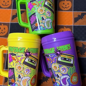 May include: Three plastic mugs in bright green, yellow, and purple, each featuring a handle and a lid. The mugs are decorated with a colorful graphic print that includes the text VINTAGE VIBES, a cassette tape, a spider, and various candy items.