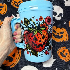 May include: A light blue plastic travel mug with a handle and a lid. The front of the mug features a graphic illustration of a red strawberry designed to look like a jack-o'-lantern with triangular eyes and a jagged mouth, surrounded by flowers.