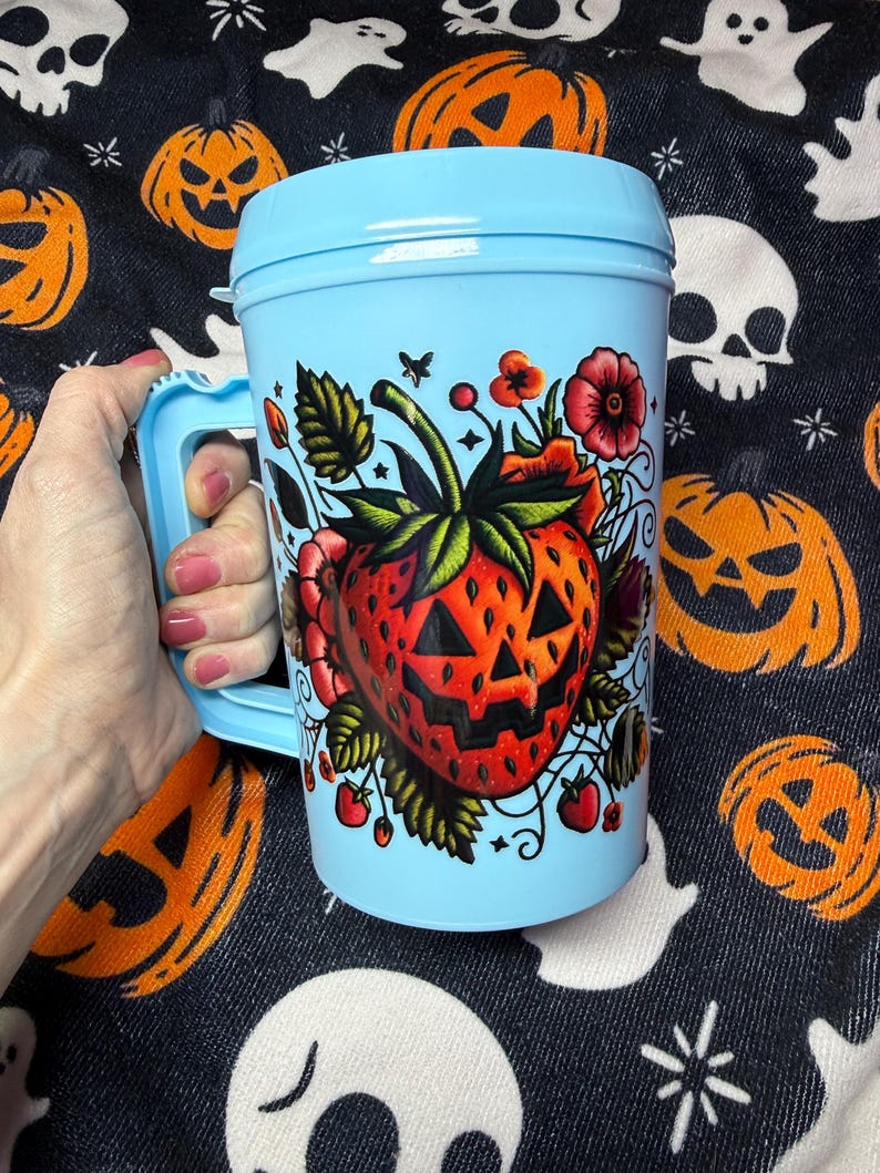 May include: A light blue plastic travel mug with a handle and a lid. The front of the mug features a graphic illustration of a red strawberry designed to look like a jack-o'-lantern with triangular eyes and a jagged mouth, surrounded by flowers.
