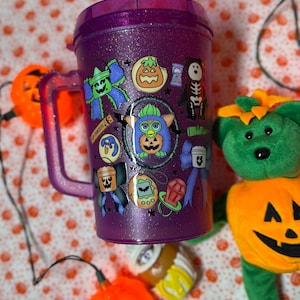 Puede incluir: Un vaso de pl&aacute;stico morado con purpurina, asa y tapa, decorado con varias pegatinas de tem&aacute;tica de Halloween que incluyen un personaje Furby central, lazos, una calabaza, un oso esqueleto, un chupete y un juguete de mascota virtual.