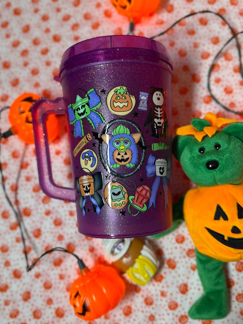 Puede incluir: Un vaso de pl&aacute;stico morado con purpurina, asa y tapa, decorado con varias pegatinas de tem&aacute;tica de Halloween que incluyen un personaje Furby central, lazos, una calabaza, un oso esqueleto, un chupete y un juguete de mascota virtual.