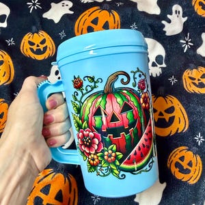May include: A light blue plastic mug with a lid and a handle, featuring a colorful graphic of a carved jack-o'-lantern pumpkin designed to look like a watermelon, surrounded by red and orange flowers, green leaves, and a slice of watermelon.