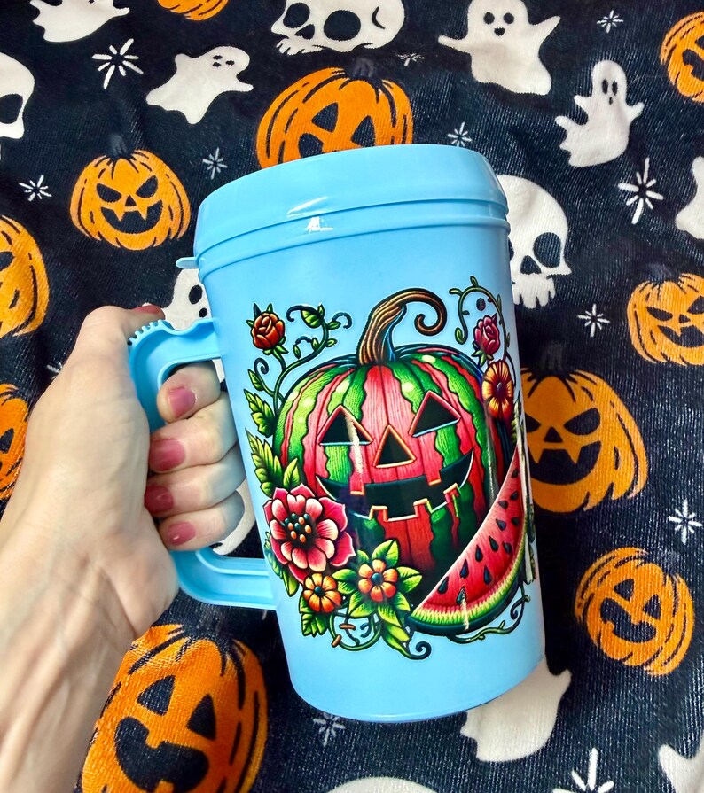 May include: A light blue plastic mug with a lid and a handle, featuring a colorful graphic of a carved jack-o'-lantern pumpkin designed to look like a watermelon, surrounded by red and orange flowers, green leaves, and a slice of watermelon.