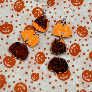 May include: A pair of dangling earrings featuring three stacked jack-o'-lantern charms each. The charms are made of reflective orange and dark copper-colored acrylic, connected by silver-toned metal jump rings and attached to silver-toned ear hooks.