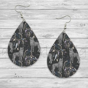 Puede incluir: Un par de pendientes en forma de lágrima con fondo negro y un patrón repetido de lobos, atrapasueños y plumas. Los pendientes son plateados y tienen cierre de gancho.