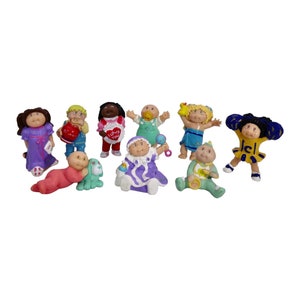 May include: A set of ten Cabbage Patch Kids figurines. The figurines are all different colors and have different outfits. Some of the figurines are holding accessories, such as a heart, a rattle, and a teddy bear.