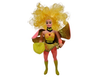 May include: A vintage action figure of a female superhero with blonde hair, wearing a pink and yellow costume with a gold belt and a yellow shield.