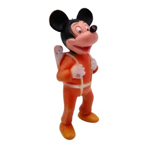 May include: A vintage plastic figurine of Mickey Mouse wearing an orange jumpsuit with a white stripe down the front and a white stripe around the waist. Mickey Mouse is wearing a black and white jetpack and has black ears and a black nose.