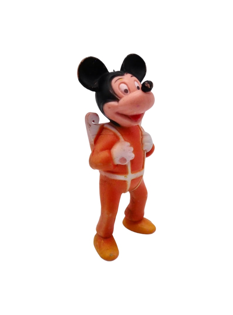May include: A vintage plastic figurine of Mickey Mouse wearing an orange jumpsuit with a white stripe down the front and a white stripe around the waist. Mickey Mouse is wearing a black and white jetpack and has black ears and a black nose.