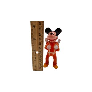 May include: A small plastic figurine of Mickey Mouse wearing an orange jumpsuit with a white harness and wings. The figurine is about 3 inches tall and is standing on a white surface.