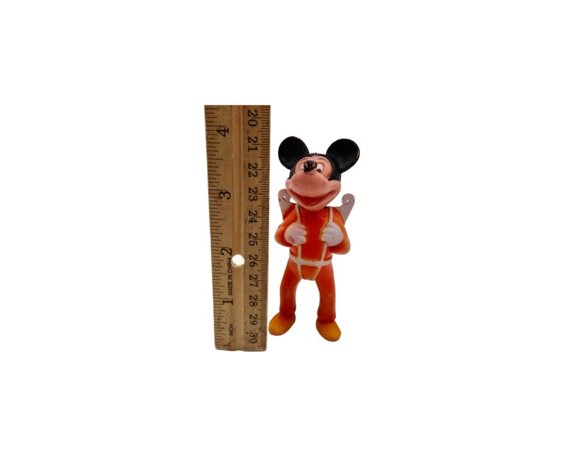 May include: A small plastic figurine of Mickey Mouse wearing an orange jumpsuit with a white harness and wings. The figurine is about 3 inches tall and is standing on a white surface.