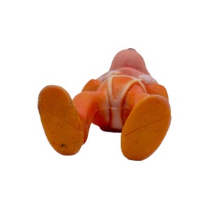 May include: Orange and white plastic toy figure of a person wearing a spacesuit. The figure is lying on its back with its legs in the air.