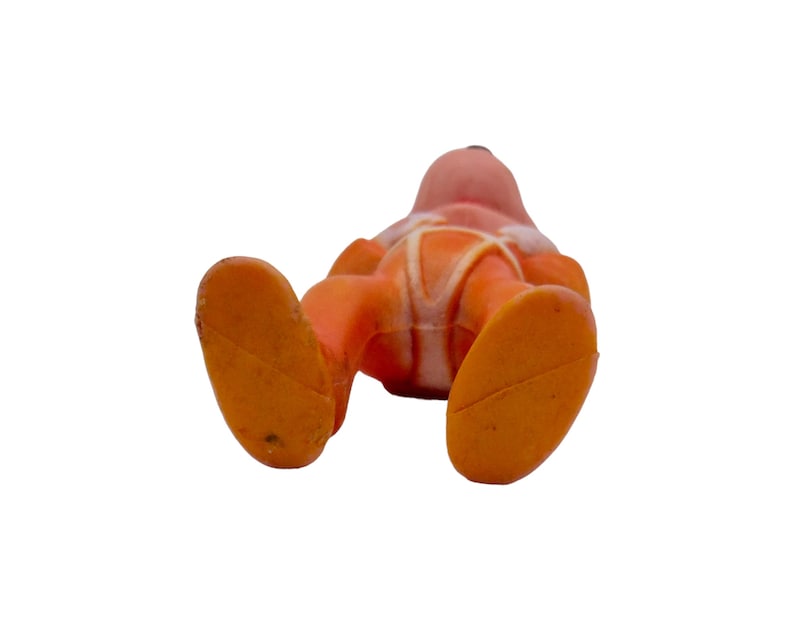 May include: Orange and white plastic toy figure of a person wearing a spacesuit. The figure is lying on its back with its legs in the air.