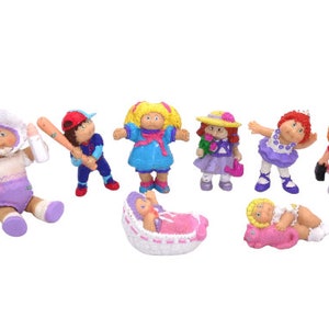 May include: A set of eight Cabbage Patch Kids figurines. The figurines are all different colors and have different outfits. There is a baby in a pink blanket, a baby in a white crib, a girl in a blue dress, a girl in a purple dress, a girl in a pink dress, a girl in a white dress, a girl in a red dress, and a boy in a blue baseball uniform.