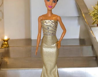 May include: A doll wearing a gold strapless gown with a sequined bodice. The doll has brown hair styled in a bun and brown eyes.