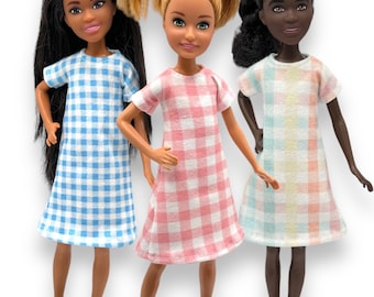 May include: Three dolls wearing gingham dresses. The doll on the left is wearing a blue and white gingham dress, the doll in the middle is wearing a pink and white gingham dress, and the doll on the right is wearing a pastel rainbow gingham dress.