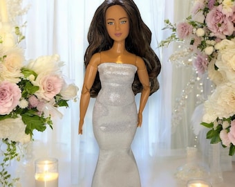 May include: A doll with long, wavy dark brown hair and tan skin, wearing a strapless, floor-length, shimmering silver gown. The doll has articulated joints at the shoulders and elbows and is standing in an upright, forward-facing position.