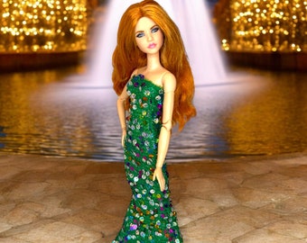 May include: A doll wearing a green sequined gown with a mermaid-style skirt. The doll has long red hair and blue eyes. The doll is standing in front of a fountain with lights in the background.