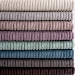 Op de afbeelding: Een stapel van acht corduroy stoffen rollen in verschillende gedempte kleuren, waaronder beige, lavendel, blauwgroen en blauw. De geribbelde textuur van de stof is duidelijk zichtbaar, waardoor een zachte, uitnodigende uitstraling ontstaat. De rollen zijn netjes opgestapeld en tonen de kleuren.