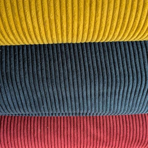 Può includere: Primo piano di tre pezzi di tessuto di velluto a coste impilati in giallo, blu navy e bordeaux. La trama a coste del tessuto è visibile, creando un'esposizione tattile e visivamente accattivante. I colori sono ricchi e saturi.