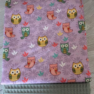 May include: A folded lavender fabric featuring a repeating pattern of cartoon owls and floral accents. Below it is a grey, textured, rectangular item. The owls are in shades of yellow, pink, and green.