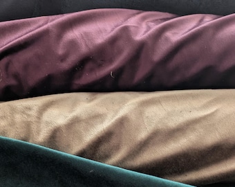 May include: Close-up of several rolls of velvet fabric in various colors. The colors include beige, navy blue, burgundy, tan, and dark green. The fabric has a soft, luxurious appearance.