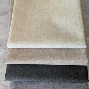 Könnte beinhalten: Ein Stapel gefalteter Stoffmuster in verschiedenen neutralen Farben. Die Muster umfassen Farbtöne von Beige, Hellbeige, Grau und Anthrazit. Der Stoff hat ein strukturiertes Aussehen, das auf ein gewebtes Material hindeutet.