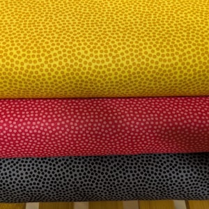 Puede incluir: Tres rollos de tela en amarillo, rojo y gris, cada uno con un patrón repetitivo de pequeños puntos espaciados. La tela está enrollada firmemente, mostrando la textura y las variaciones de color. La tela amarilla está en la parte superior, seguida de la roja y luego la gris.